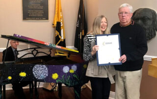 Two people holding a citation by a colorful painted piano with a pianist.